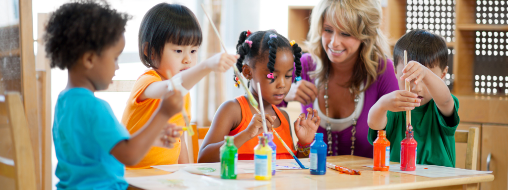 A carer smiles as 4 young children paint around a table together