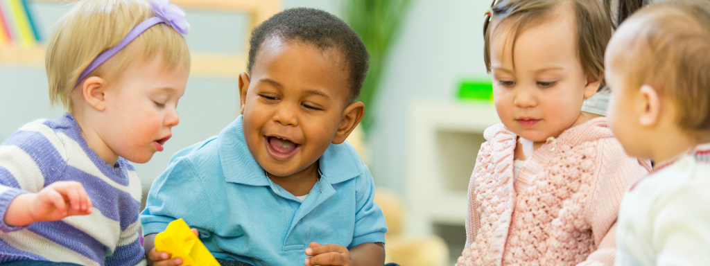 Four toddlers sit together, one is holding a yellow brick