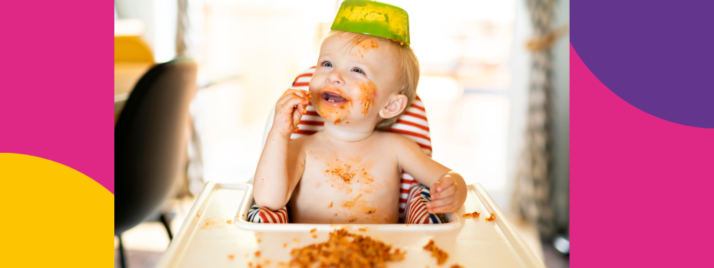 A baby eats spaghetti, with a bowl on their head